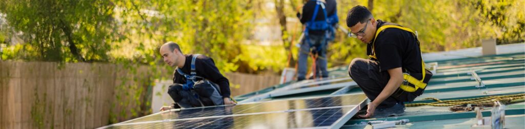 Two workers, wearing safety harnesses and glasses, installing solar panels on a green metal roof outdoors on a sunny day.